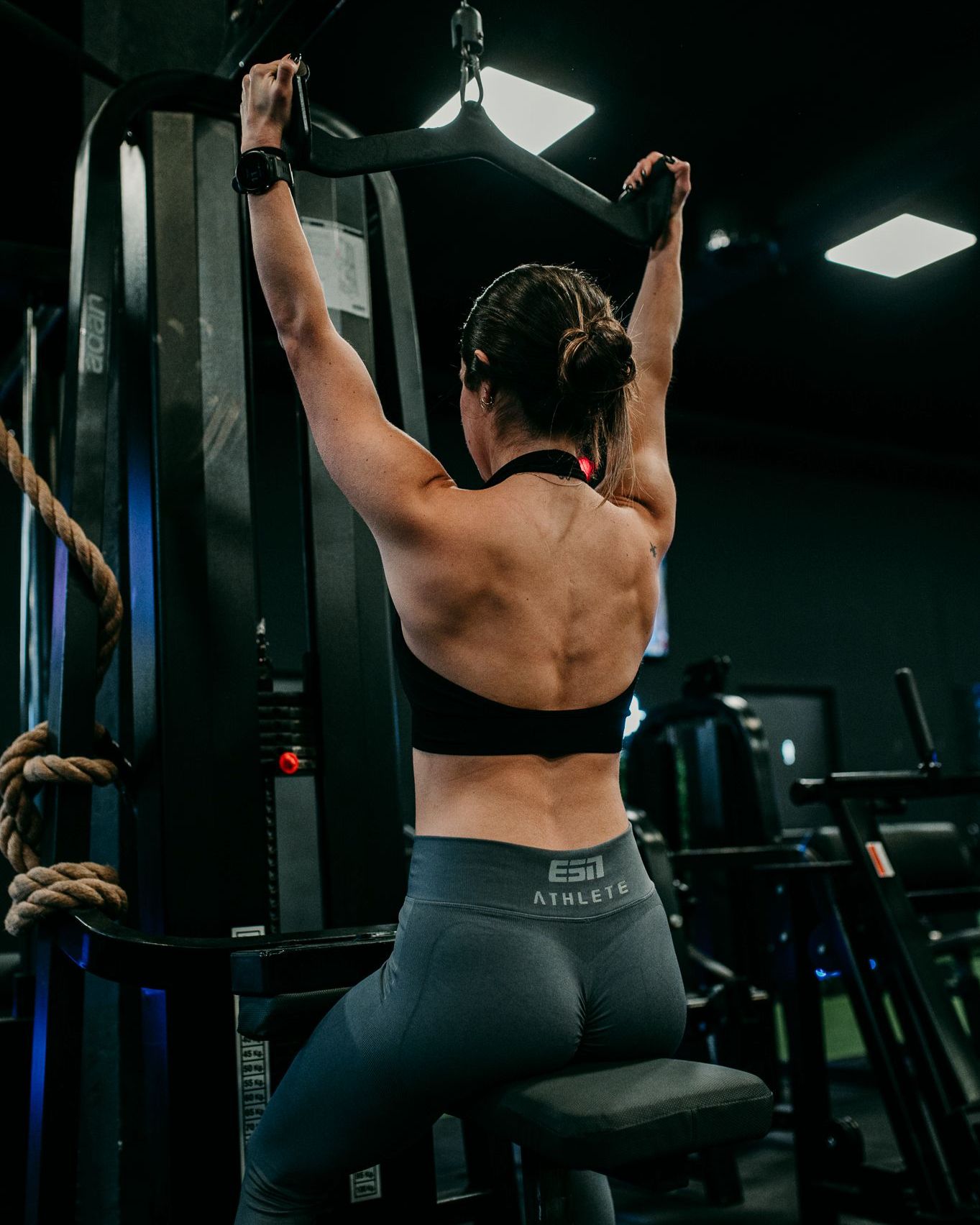 La entrenadora usa una máquina de polea en el gimnasio con top negro y mallas grises.