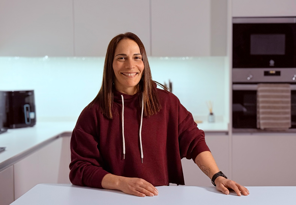 Entrenadora con un suéter burdeos de pie en la cocina con una gran sonrisa.