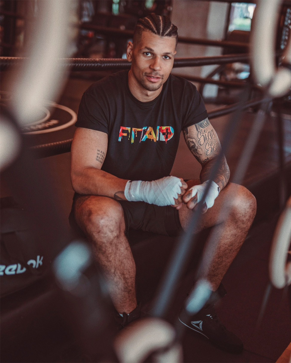 De coach met gevlochten haar zit in een boksring met zwachtels om zijn handen en draagt een zwart T-shirt.