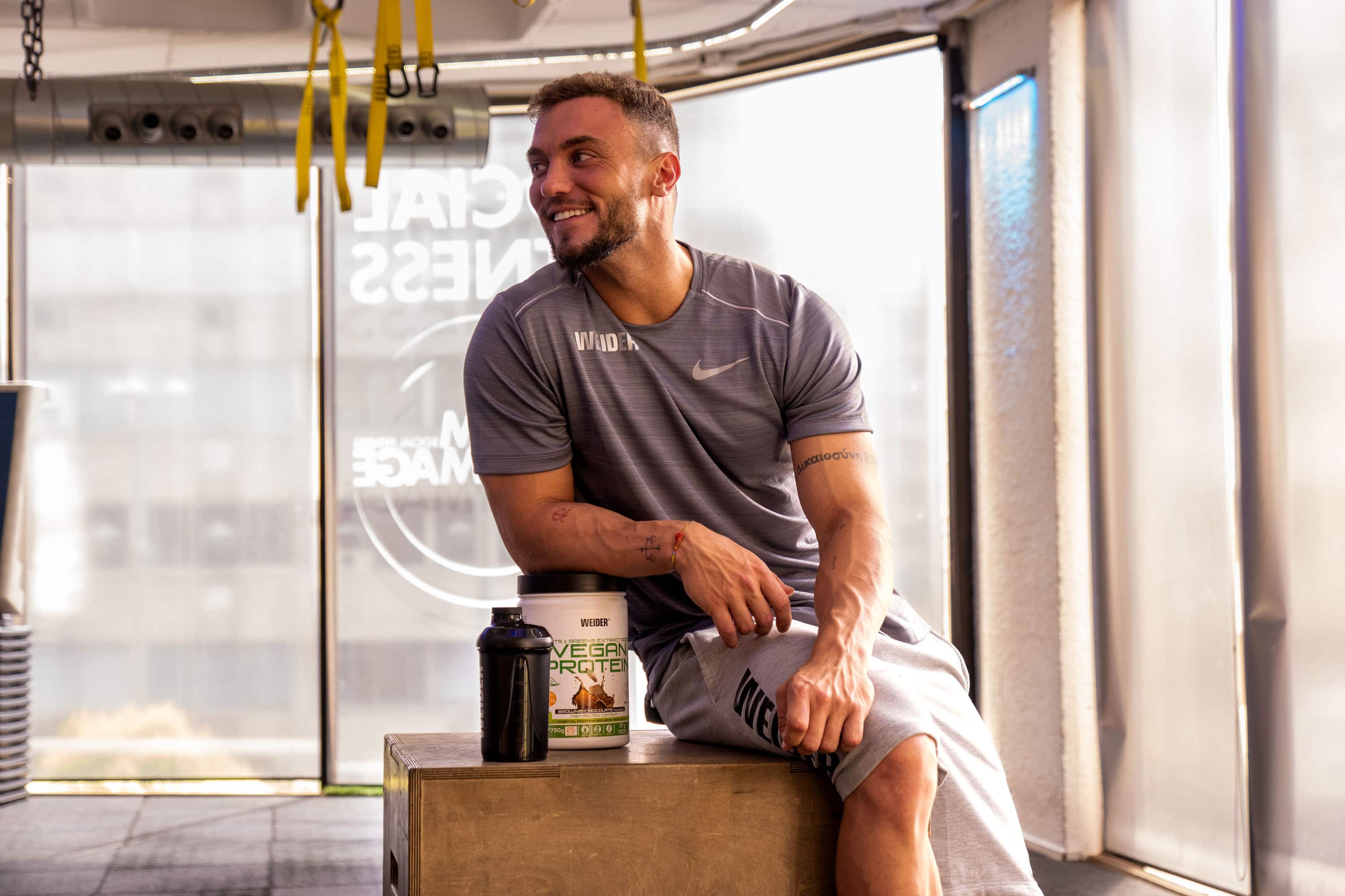 Coach Alberto sonriendo y apoyando el brazo en un envase de proteína vegana en polvo.